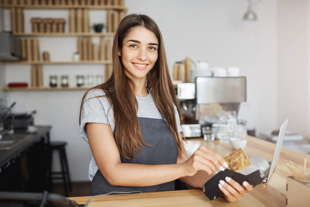 Agência Simi empregada-feminina-da-cafeteria-usando-um-leitor-de-cartao-de-credito-para-cobrar-cliente-parecendo-feliz-sorrindo-para-camera