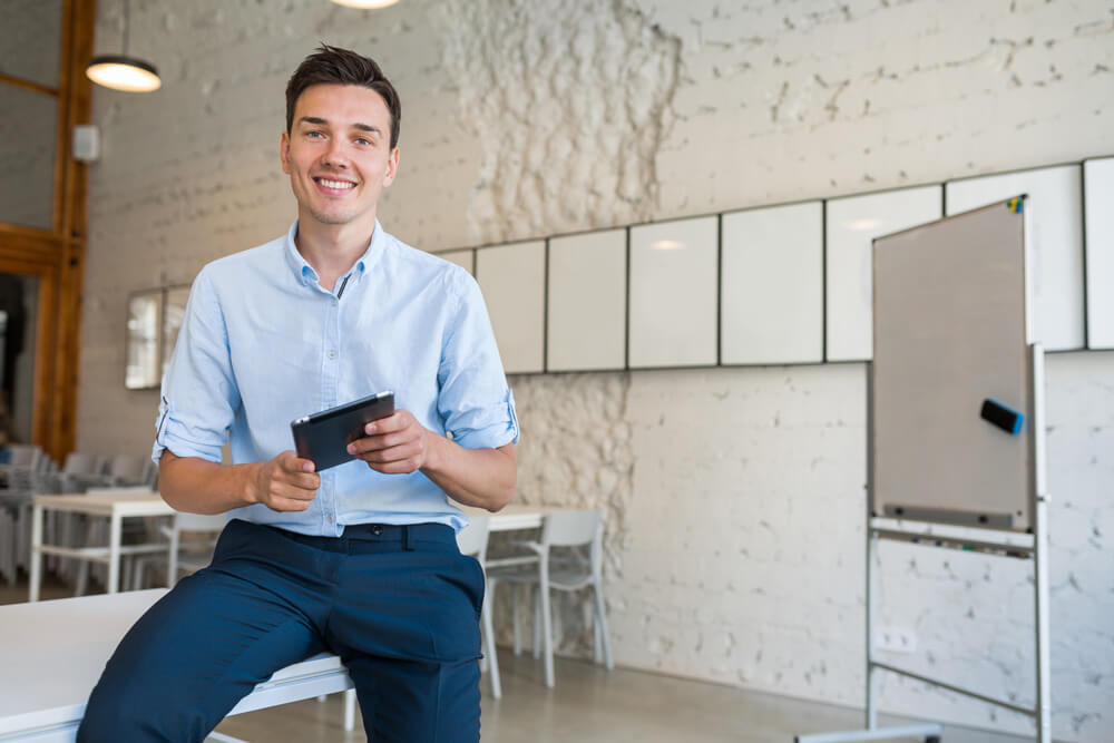 Agência Simi feliz-jovem-elegante-e-sorridente-no-escritorio-de-co-trabalho-freelancer-de-inicializacao-segurando-usando-tablet