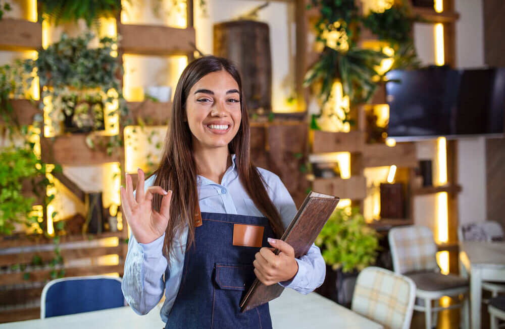 Agência Simi feliz-linda-garconete-sorridente-usando-avental-segurando-um-menu-de-pasta-em-um-restaurante-olhando-para-camera-em-pe-no-cafe-aconchegante-bom-servico