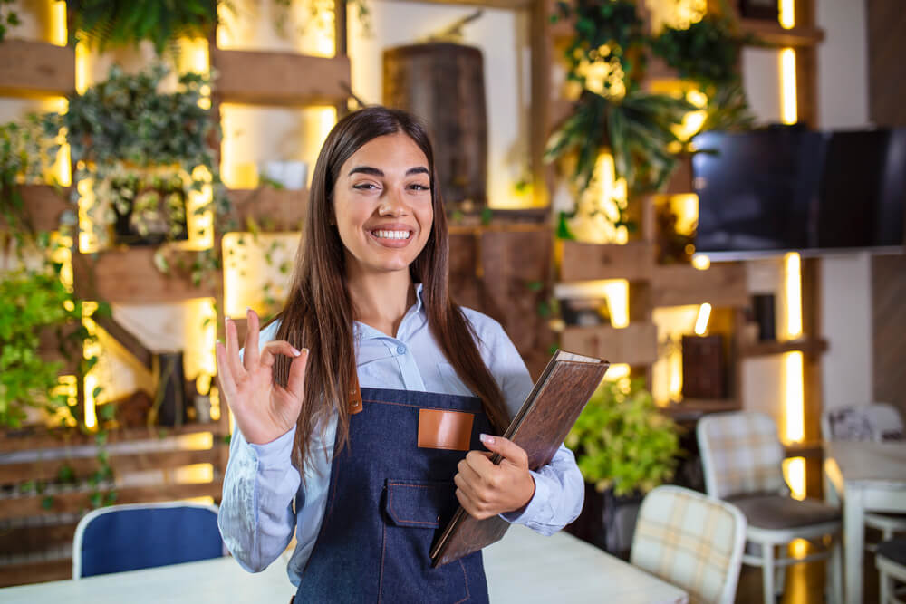 Agência Simi feliz-linda-garconete-sorridente-usando-avental-segurando-um-menu-de-pasta-em-um-restaurante-olhando-para-camera-em-pe-no-cafe-aconchegante-bom-servico